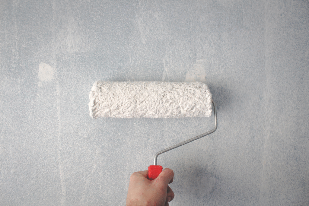 Hand holding a paint roller applying primer on a textured wall, illustrating preparation for painting in an interior house painting project.