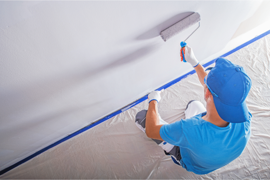 Person painting a textured wall with a roller, wearing gloves and a blue cap, on a drop cloth with painter's tape along the edge.