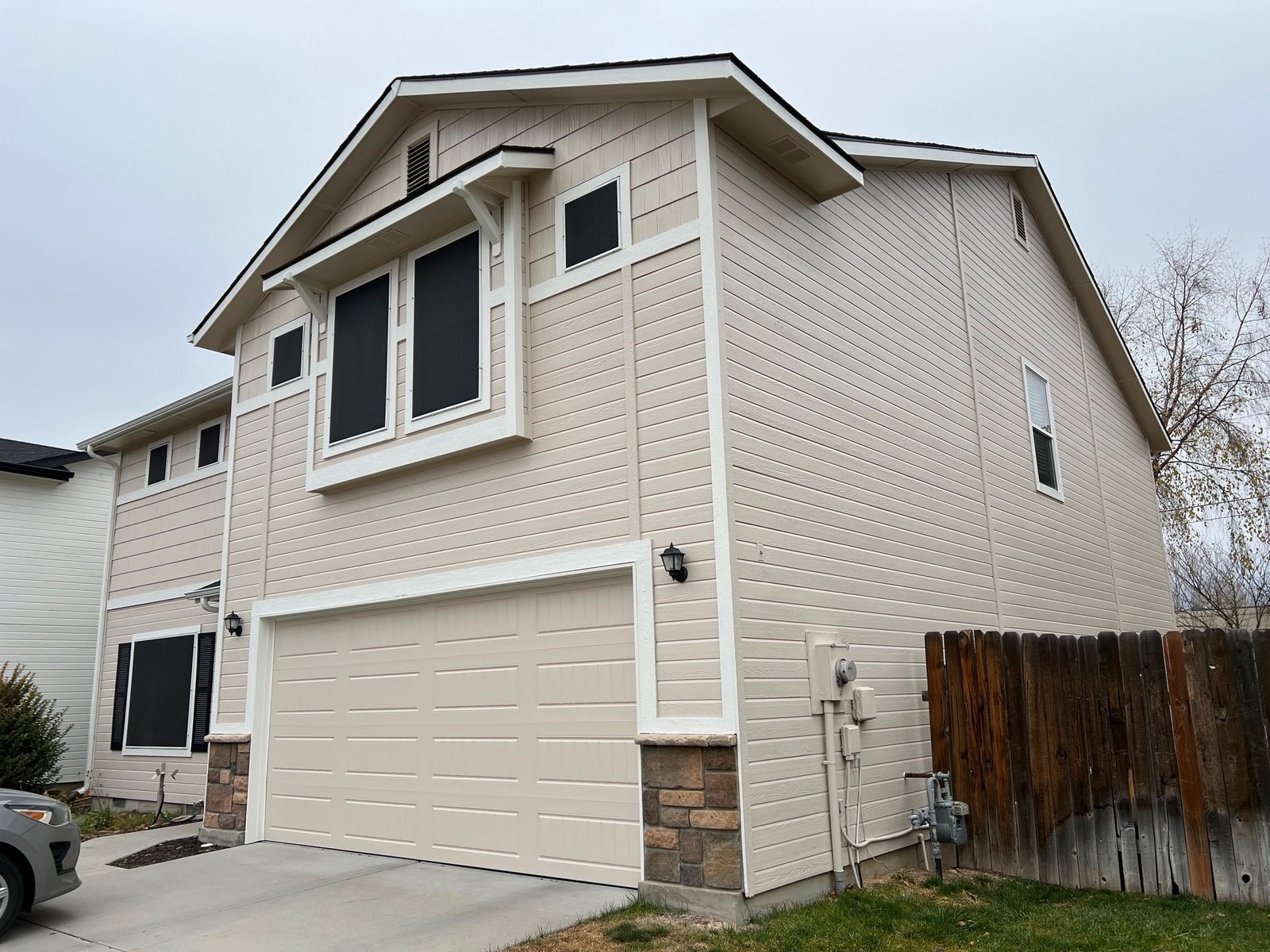 Exterior view of a two-story house with light beige siding, black window screens, and a garage door, illustrating proper surface preparation for painting in spring weather conditions.