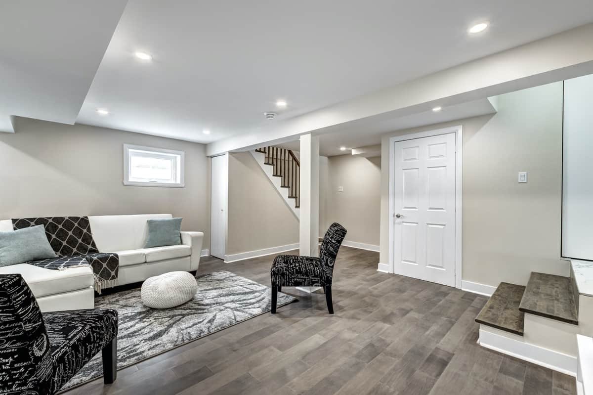 Modern interior of a basement living space featuring a white sofa with decorative pillows, a patterned area rug, black accent chairs, and wooden stairs, emphasizing a clean and inviting atmosphere for potential interior painting services in Nampa, ID.